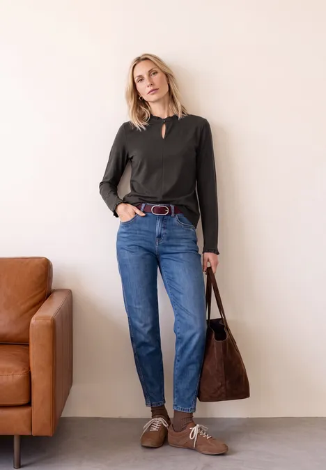 Basic Shirt met ruches en een uitsnijding black coffee Frau steht vor weißer Wand in schwarzem Langarmshirt, blauen Jeans, braunen Schuhen und großer brauner Tasche.