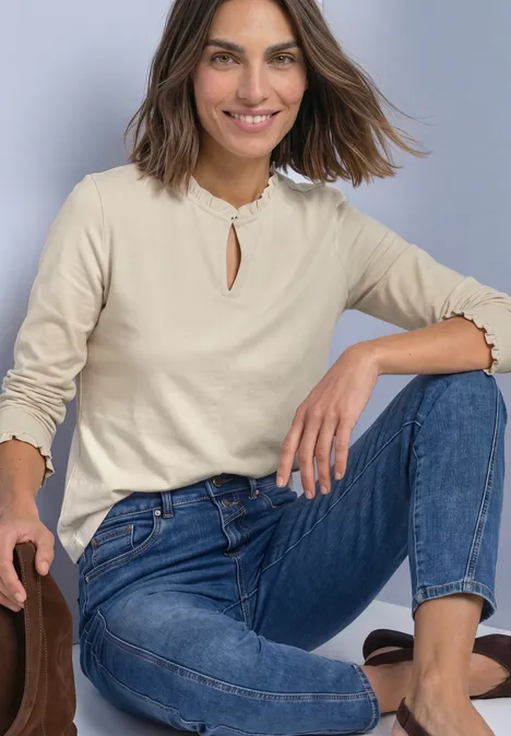 Frau kniet lässig, trägt beiges Langarmshirt mit Rüschen und blaue Jeans.