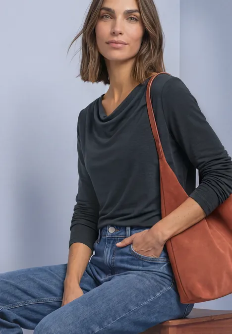 Vrouw in zwart shirt met watervalhals, jeans en bruine schoudertas, zittend met een lichte glimlach.