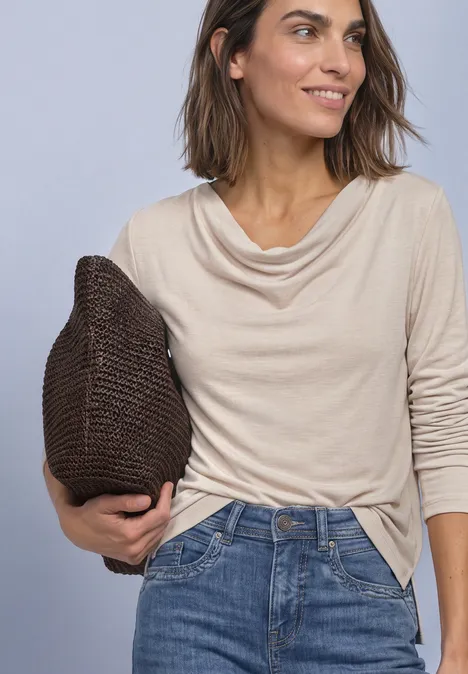 Vrouw glimlachend in beige shirt met watervalhals, blauwe jeans en bruin gebreid tasje.