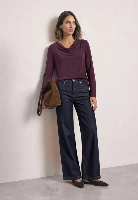 Vrouw in paarse coltrui, wijde jeans, bruine tas en platte loafers voor een effen muur.