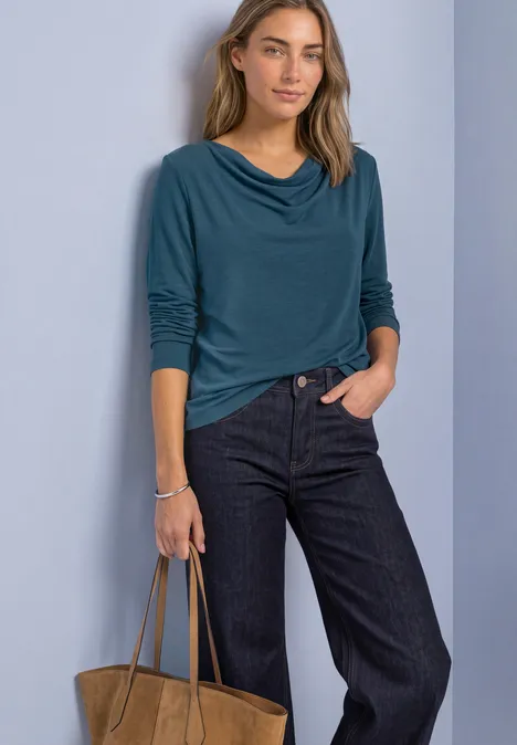 Vrouw draagt een teal top met watervalnek, wijde jeans en houdt een bruine tas vast voor een lichte achtergrond.