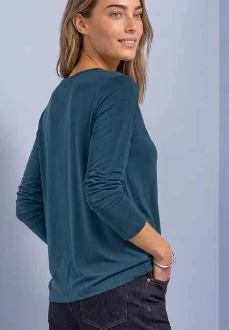Vrouw van achteren in een blauwgroen shirt met lange mouwen, rechterhand in haar spijkerbroekzak