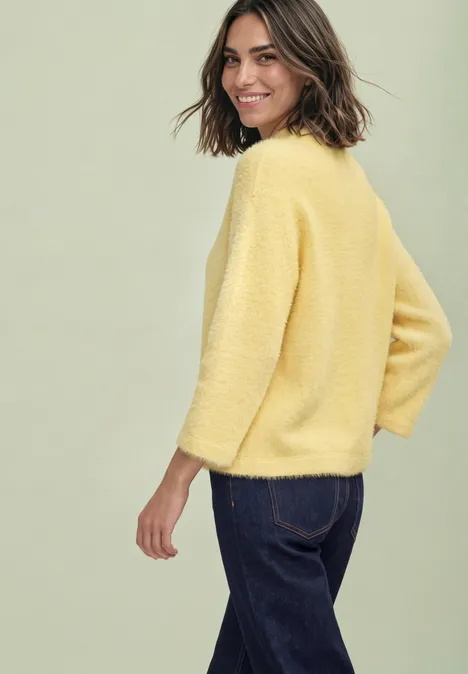 Junge Frau in gelbem Pullover und blauer Jeans, lächelnd seitlich in die Kamera blickend.