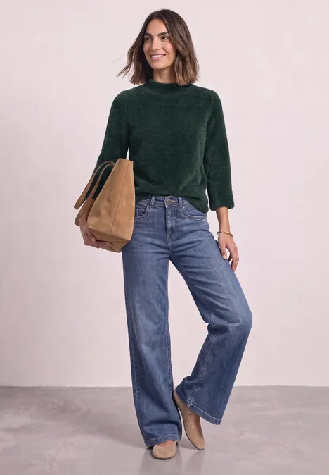 3/4-Arm Shirt mit Turtle Neck stormy green Frau in grüner Bluse und weitem Jeans, hält eine braune Tasche, vor hellem Hintergrund.