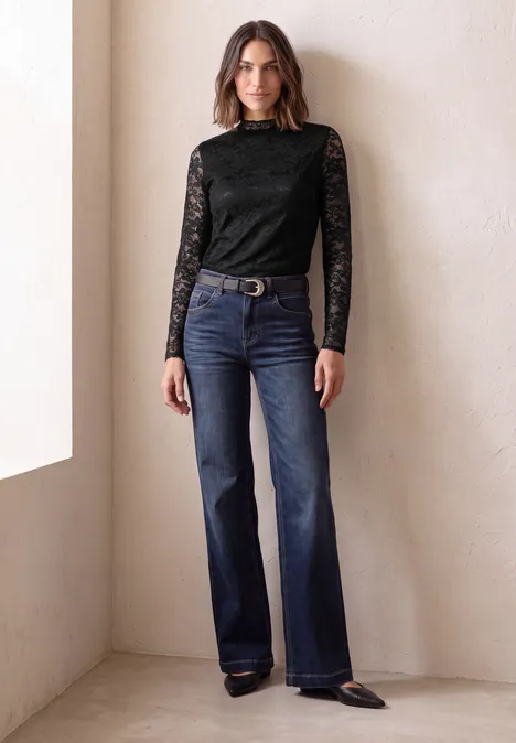 Vrouw in zwarte kanten top en wijde blauwe jeans staat voor een lichte muur.