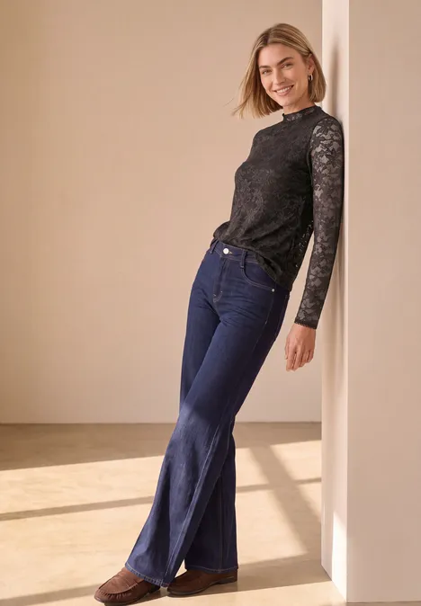 Jonge vrouw leunend tegen een muur in een lichte kamer, glimlachend in een kanten top en wijde jeans.