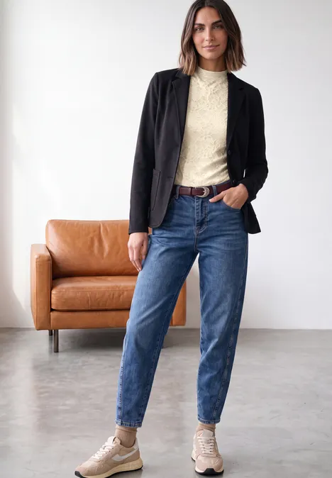 Een vrouw in een zwarte jas, crèmekleurige trui, blauwe jeans en beige laarzen staat in een modern interieur.