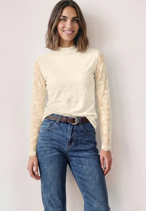 Vrouw met bruin haar in crèmekleurige kanten top en blauwe jeans, glimlachend voor een lichte achtergrond.