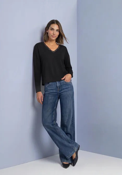T-shirt à manches longues, col en V et détail en dentelle Black Frau steht vor neutralem Hintergrund, trägt schwarze V-Ausschnitt-Bluse und weite blaue Jeans mit schwarzen Schuhen.