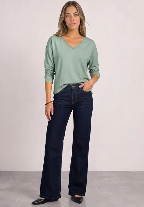 Langarm Shirt mit V-Neck und Spitzendetail eucalyptus green Eine Frau in hellgrünem Pullover und dunkelblauen Schlagjeans steht lässig.