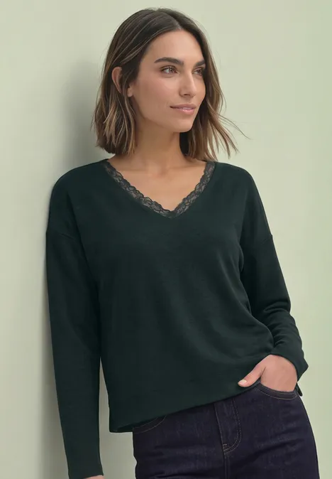 T-shirt à manches longues, col en V et détail en dentelle stormy green Frau in schwarzem Langarmshirt mit Spitzen-V-Ausschnitt und Jeans, steht entspannt mit Hand in Tasche.
