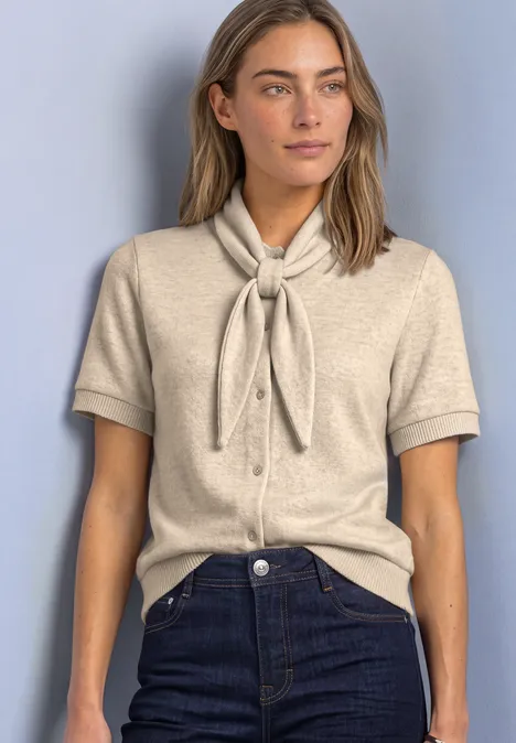 Frau in beiger Kurzarm-Strickbluse mit Schleifenkragen, dunkelblaue Jeans, vor hellblauem Hintergrund.
