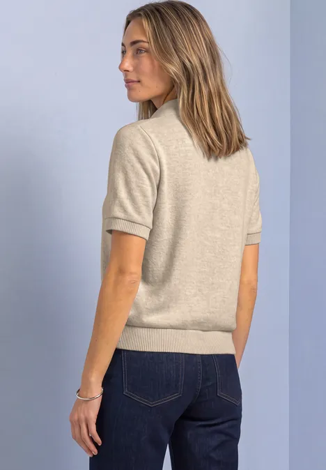 Frau in beigeem Kurzarm-Pullover und blauen Jeans im seitlichen Profil vor hellem Hintergrund.