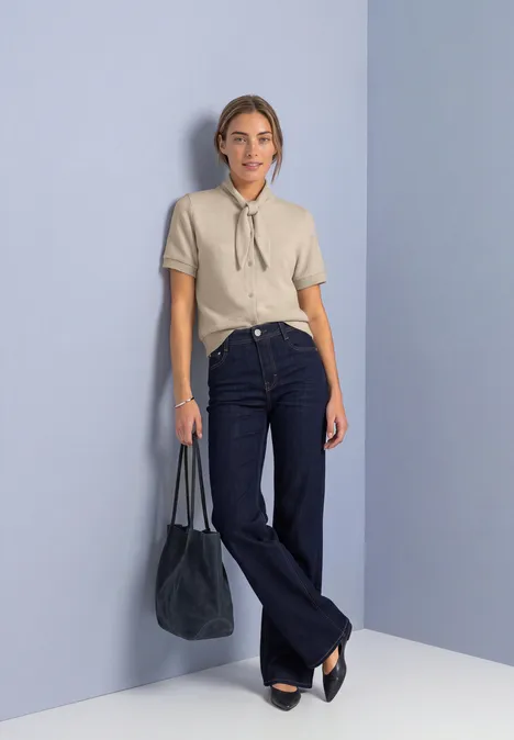 Frau in beiger Bluse mit Schluppe, dunkelblaue Schlagjeans und schwarzer Tasche vor pastellfarbener Wand.