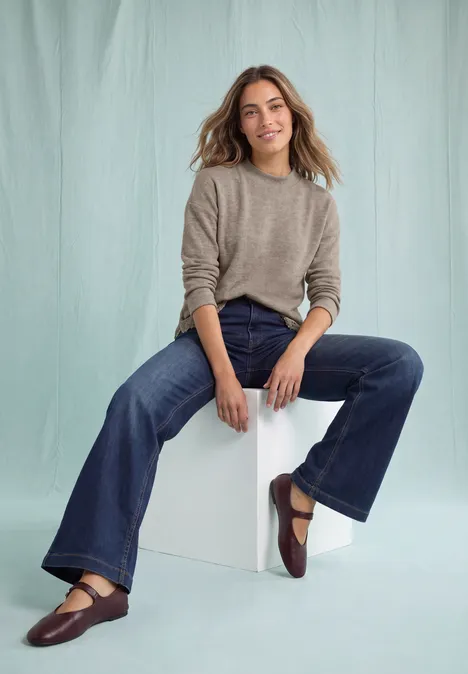 Langarm Shirt mit Rundhals und Spitzenborte sanded amber mel. Eine lächelnde Frau sitzt auf weißem Würfel, trägt graues Strickshirt, ausgestellte Jeans und braune Ballerinas.