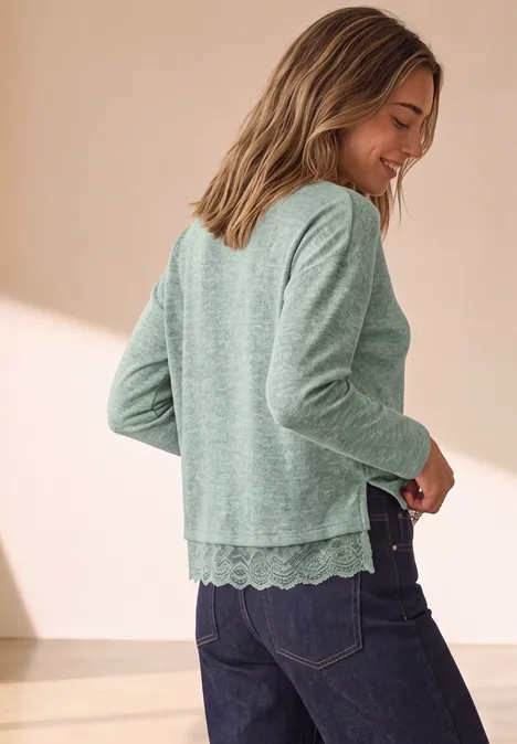 Langarm Shirt mit Rundhals und Spitzenborte eucalyptus green mel. Frau trägt grünen Pullover mit Spitzenbesatz und Jeans, steht seitlich und lächelt.