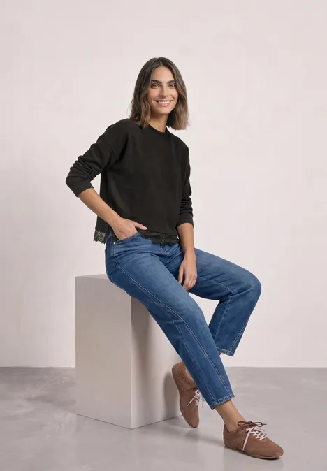 Langarm Shirt mit Rundhals und Spitzenborte black coffee mel. Frau sitzt auf weißem Würfel, trägt schwarzen Pullover, blaue Jeans und braune Schuhe, lächelt leicht.