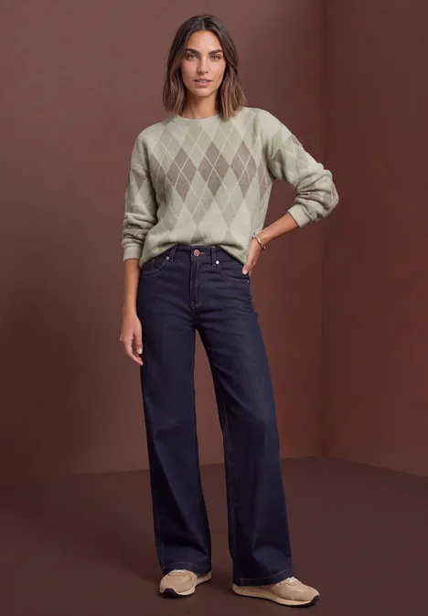T-shirt à manches longues, col rond et motif à carreaux eucalyptus green Frau in grünem Rautenmuster-Pullover, weiten Jeans und hellen Slippers vor braunem Hintergrund.