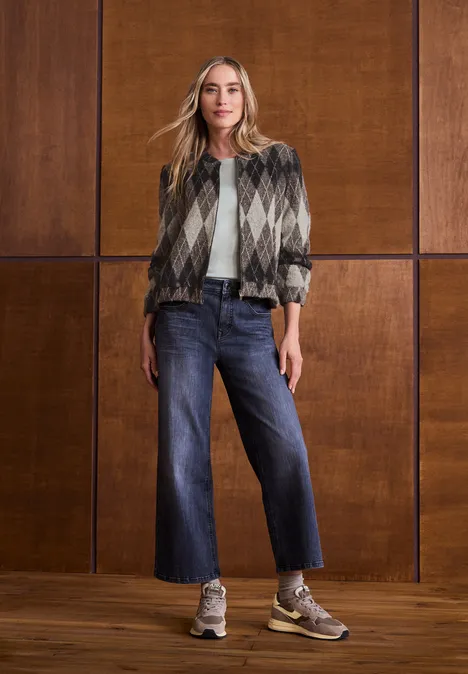 Blouson à manches longues à carreaux black coffee Femme portant une veste à carreaux, un t-shirt blanc, jean large et baskets devant un mur en bois.