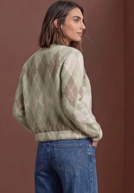 Langarm Blouson mit Karomuster eucalyptus green Frau in Rauten-Strickpullover in Grün und Creme, Jeans, steht seitlich vor braunem Hintergrund.