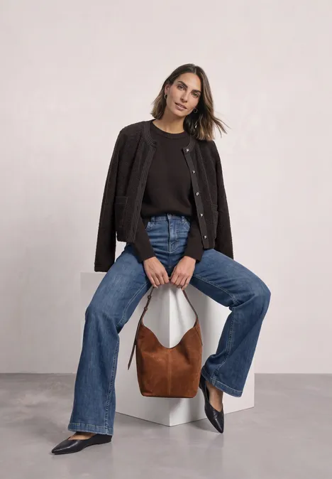 Teddy-Jacke mit Strukturdetails black coffee Frau sitzt auf weißem Sockel, trägt schwarzen Cardigan, Jeans und schwarze Schuhe, braune Tasche neben ihr.