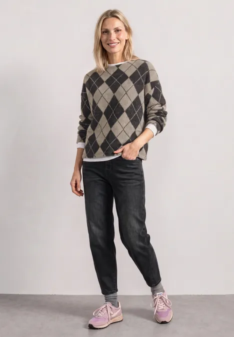 Vrouw lacht in een geruite trui, zwarte jeans en roze sneakers, staand voor een neutrale achtergrond.