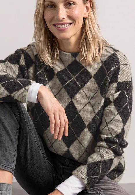 Een glimlachende vrouw in een geruite trui en jeans zit ontspannen, met ruitpatroon.