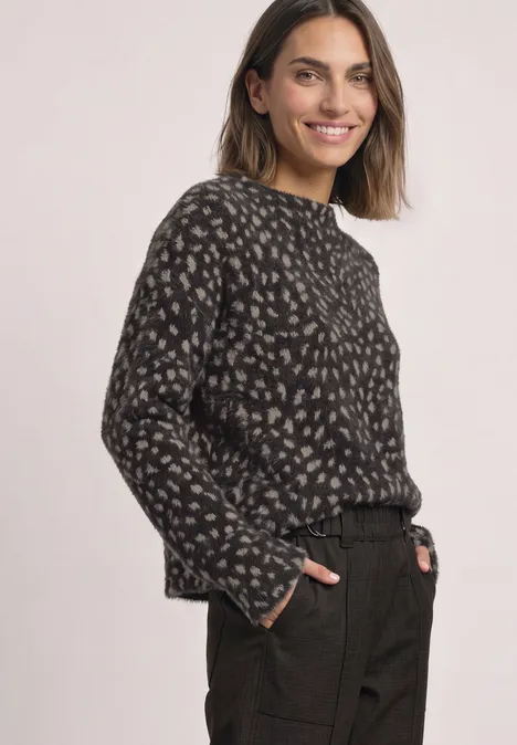 Lachende vrouw in donkere, gemêleerde trui met licht patroon en zwarte broek voor lichte achtergrond.