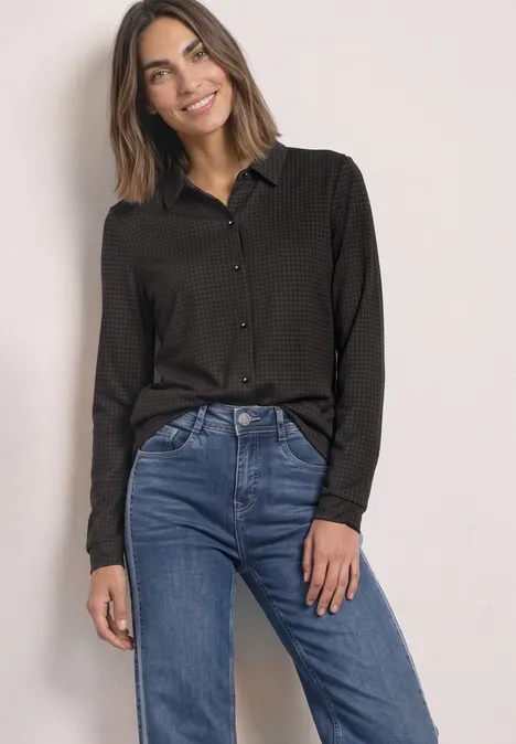 Vrouw in zwarte blouse en blauwe spijkerbroek tegen neutrale achtergrond, glimlacht.