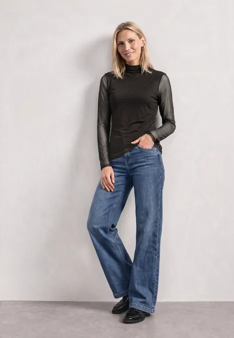 T-shirt en mesh à col roulé froncé black coffee Frau vor neutralem Hintergrund trägt schwarzen Langarmtop, weite blaue Jeans und schwarze Schuhe.