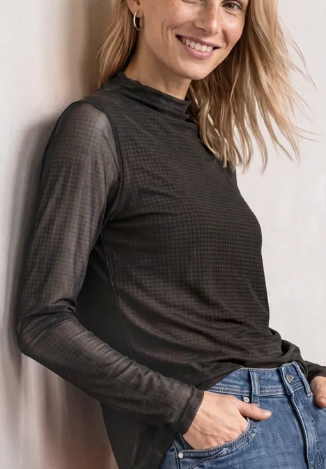 T-shirt en mesh à col roulé froncé black coffee Frau lehnt an Wand, trägt schwarzen transparenten Langarmpullover und Jeans, lächelt leicht.
