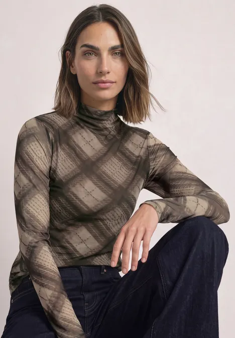 Frau sitzt vor hellem Hintergrund, trägt transparent karierten Rollkragenpullover und dunkle Jeans.
