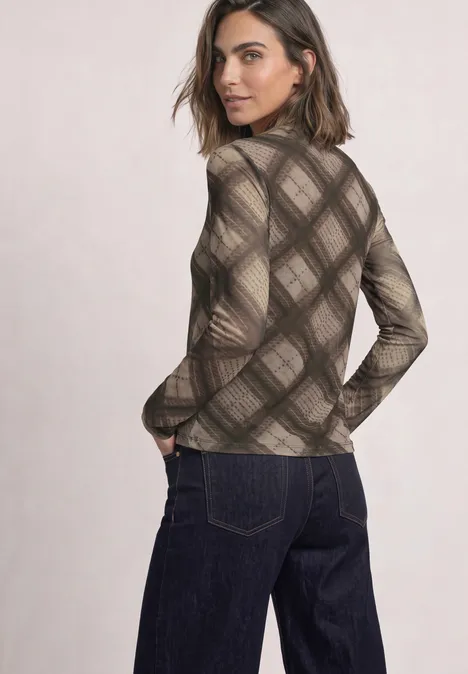 Eine Frau im braunen, karierten Rollkragenpullover und dunklen Jeans, sie blickt über die Schulter.