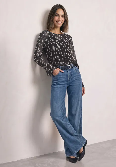 Shirt aus Mesh mit langen Volant-Ärmeln Black Eine Frau trägt ein schwarzes, gemustertes Langarmshirt und weite Jeans, steht vor heller Wand.