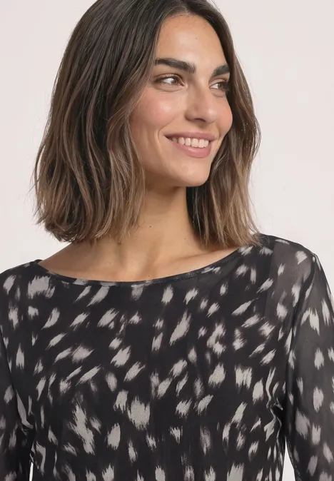Shirt aus Mesh mit langen Volant-Ärmeln Black Frau mit schulterlangem dunklem Haar, seitlich lächelnd, trägt schwarzes, gemustertes Oberteil.