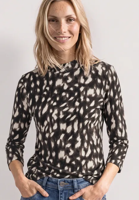 Eine lächelnde Frau im schwarz-beigen, gepunkteten Langarmshirt und Jeans vor weißem Hintergrund.