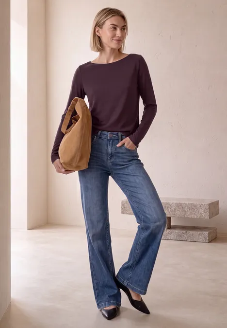 Vrouw in paars langarmshirt, rechte blauwe jeans en zwarte ballerina’s, draagt een bruine tas.
