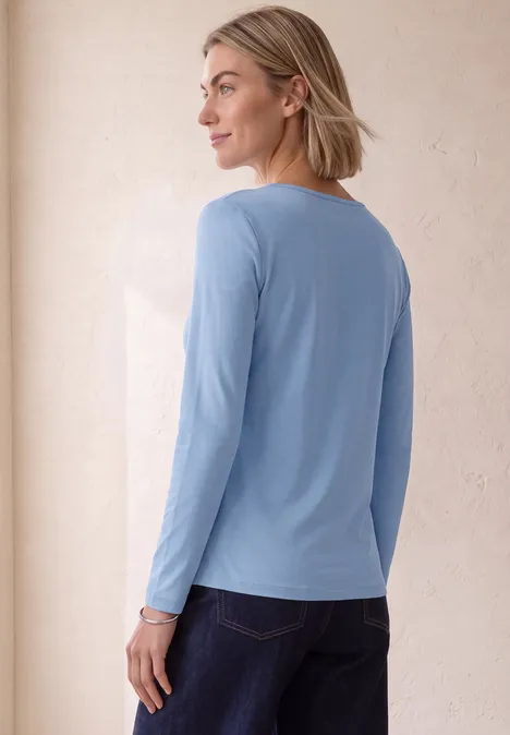 Frau mit schulterlangem blonden Haar trägt ein hellblaues Langarmshirt und blickt seitlich über die Schulter.