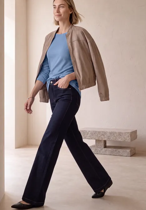 Frau im blauen Top und weiten Jeans mit hellbrauner Jacke über den Schultern vor minimalistischem Hintergrund