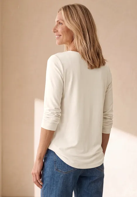 Een vrouw van opzij draagt een wit langarmshirt en een blauwe spijkerbroek tegen een lichte achtergrond.
