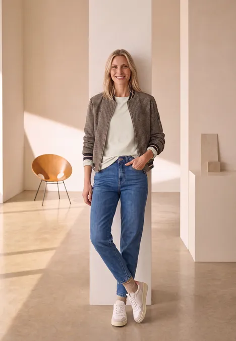 Een lachende vrouw in jeans en sneakers leunt tegen een zuil in een lichte, moderne ruimte.