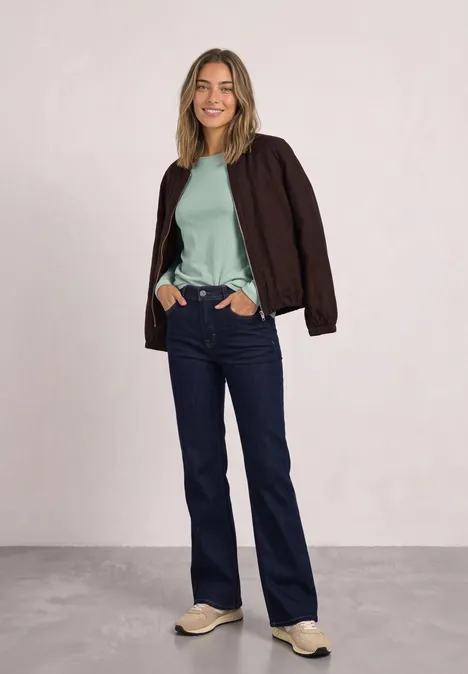 T-shirt basique à col bateau eucalyptus green Frau in Jeans, mintgrünem Shirt, brauner Jacke und Sneakern steht lächelnd mit Händen in den Taschen vor neutralem Hintergrund.