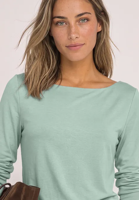 T-shirt basique à col bateau eucalyptus green Frau im mintgrünen Langarmshirt, hält eine braune Tasche vor hellem Hintergrund.
