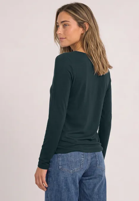 T-shirt basique à col bateau stormy green Junge Frau im Profil mit dunkelgrünem Langarmshirt und blauen Jeans vor hellem Hintergrund.