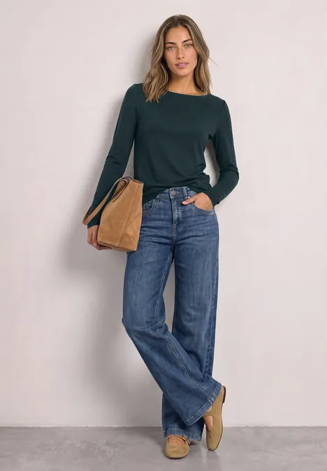 T-shirt basique à col bateau stormy green Frau in dunkelgrünem Pullover, weiten Jeans und beiger Tasche vor neutraler Wand.