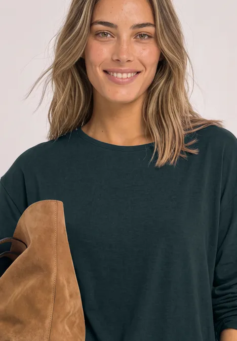 T-shirt basique à col bateau stormy green Eine lächelnde Frau mit braunen Haaren trägt ein dunkles Oberteil und eine braune Wildledertasche.