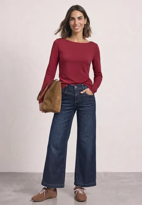 Vrouw in rood shirt met lange mouwen, wijd donkerblauw spijkerbroek, bruine schoenen en tas tegen lichte achtergrond.