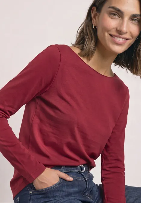 Glimlachende vrouw in een rood shirt met lange mouwen en jeans, hand in de zak.