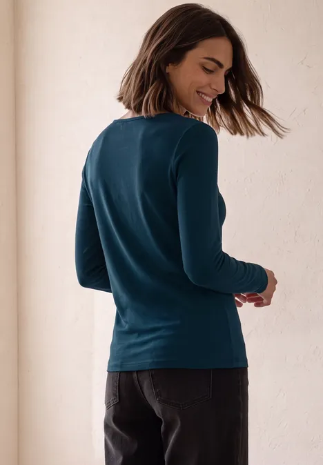 Vrouw in donkerblauw shirt met lange mouwen en zwarte broek, half van opzij, glimlachend.